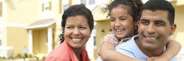 A smiling mother, father and daughter in front of a home.